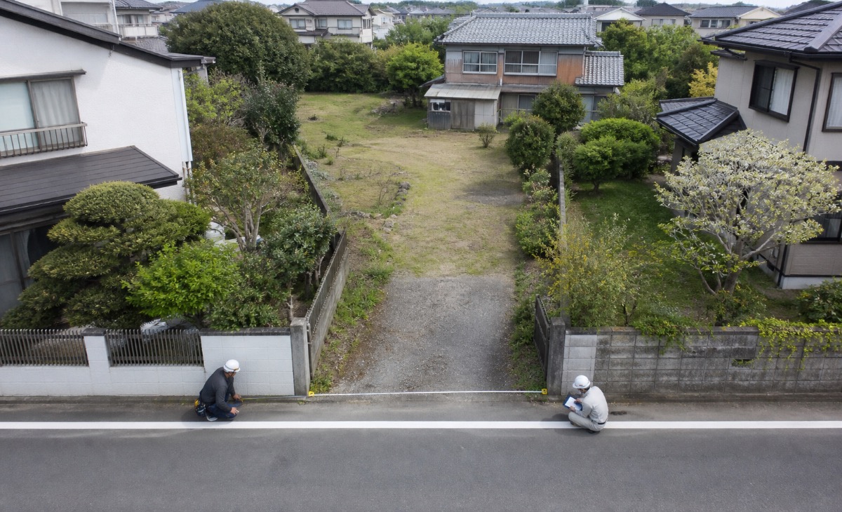 公道から細い進入路と奥の広い敷地を見渡し、入口幅を測定する作業イメージ
